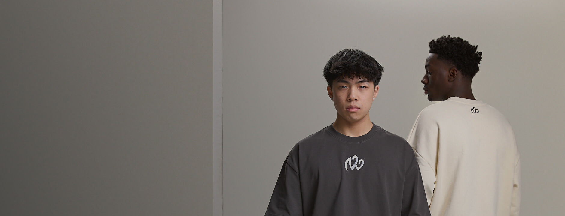 Charcoal crewneck with white chest logo; cream crewneck shows back neck print — clean studio streetwear hero.